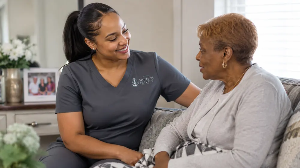 A caregiver and an older woman talking together on a sofa in a bright living room.